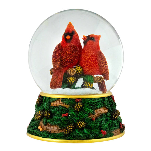 Snow globe with two cardinals on a branch inside, surrounded by pinecones and leaves.