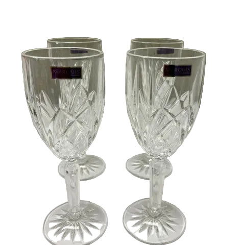 Four clear cut-crystal wine glasses with faceted stems and round starburst bases, arranged on a white surface against a plain beige background. Small purple brand tags are visible near the rims.
