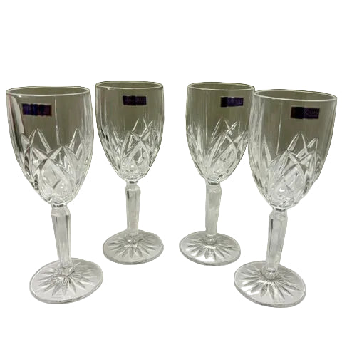 Four clear cut-crystal wine glasses with faceted stems and circular bases, arranged on a white cloth against a beige wall.